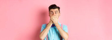 Ağzı ellerle ve şaşı gözlerle kaplayan komik genç adam, palyaço suratları yapıyor, pembe arka planda aptal aptal duruyor, insanları eğlendiriyor..