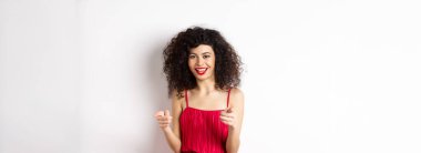 Kırmızı elbiseli güzel kadın seni tebrik ediyor, kamerayı işaret ediyor ve gülümsüyor, beyaz arka planda duruyor..