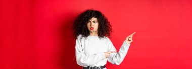 Hayal kırıklığına uğramış komik kadın, somurtuyor ve şikayet ediyor, boş alanı işaret ediyor ve tartışıyor, hoşnutsuz görünüyor, kırmızı arka plan.