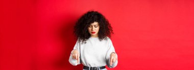 Kıvırcık saçlı endişeli genç bir kadın, yere bakıyor ve endişeli, tereddütlü bir yüzle, kırmızı arka planda duruyor..