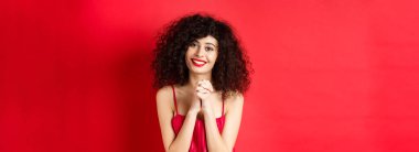 Kırmızı elbiseli ve makyajlı güzel kıvırcık saçlı bir kadın, teşekkür ediyor, kamera karşısında mutlu ve gülümsüyor..