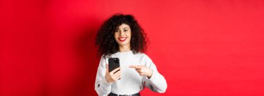 Akıllı telefonlu neşeli bir bayan. Telefonunu işaret ediyor. Tatmin olmuş bir şekilde gülümsüyor..
