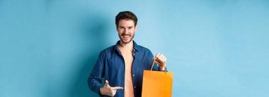 Gülümseyen beyaz adam mavi arka planda duran turuncu alışveriş çantasına işaret ediyor..