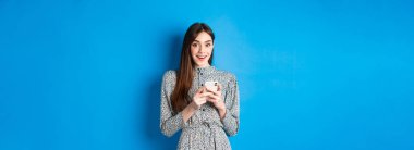 Uzun, doğal saçları olan, elbise giyen, akıllı telefon kullanan ve mutlu görünen, mavi arka planda duran tatlı bir kız..