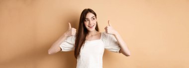 Gülümseyen hayalperest kız sol üst köşeye bakıyor ve reklamlarda baş parmak gösteriyor, ürün öneriyor, afişi övüyor, bej arka planda duruyor..