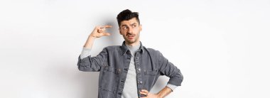 Düşünceli genç adam yana bakıyor ve düşünüyor, küçük bir nesne gösteriyor, küçük ya da küçük bir şeyi düşünüyor, beyaz arka planda duruyor..