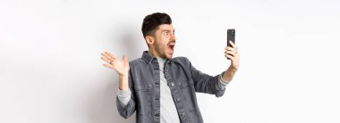 Şaşırmış ve kafası karışmış bir adam akıllı telefon ekranına bakıyor, hayal kırıklığından bağırıyor, beyaz arka planda duruyor..