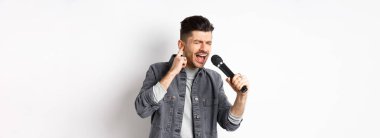 Yakışıklı şarkıcı, yüksek notaya ulaşmaya çalışıyor, mikrofonla şarkı söylüyor ve bir kulağını kapatıp beyaz arka planda duruyor..