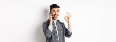 Heyecanlı genç adam telefonda konuşuyor ve iyi bir hareket gösteriyor, gülümsüyor, beyaz arka plana karşı duruyor..