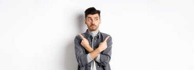 Kararsız yakışıklı adam parmaklarını yana çeviriyor, yana bakıyor ve seçimler yapıyor, iki varyantı gösteriyor, beyaz arka planda kararsız duruyor..