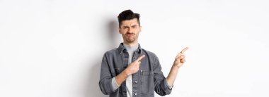 Şüpheci ve hayal kırıklığına uğramış genç adam solu işaret ediyor, reddedilerek kafasını sallıyor, kötü ürünlerden hoşlanmıyor, beyaz arka planda duruyor..