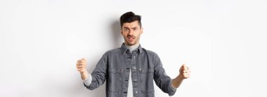 Şüpheci genç adam büyük bir nesne gösteriyor ve somurtuyor, beyaz arka planda isteksiz duruyor..