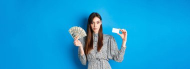 Alışverişe. Dolar banknotları, para ve plastik kredi kartı olan zarif, küstah kadın, kameraya bakıyor, mavi arka plan..