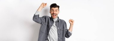 Heyecanlı adam sevinçten bağırıyor, ödül kazanıyor ve seviniyor, ellerini kaldırıyor ve evet diye bağırıyor tatmin olmuş yüzle, zaferle, hedefe ulaşıyor, beyaz arka planda duruyor..