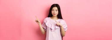 Arsız Asyalı kız ilginç bir reklam veriyor, parmaklarını logoya doğrultuyor ve kurnazca gülümsüyor, iyi bir anlaşma öneriyor, pembe arka plan..