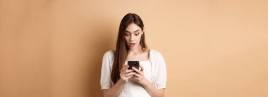 Kadın akıllı telefon ekranına şaşırmış görünüyor, cep telefonundan şok edici bir mesaj okuyor, bej arka planda şaşkın şaşkın duruyor..
