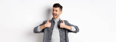 Bıyıklı mutlu genç adam göz kırpıyor ve baş parmağını kaldırıyor, bir şey tavsiye ediyor, iyi bir şey övüyor, iltifat ediyor, beyaz arka planda duruyor..