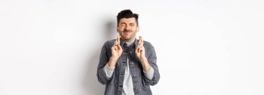 Umut dolu genç adam, gözleri kapalı ve parmakları kapalı, dilek diliyor, rüya için tanrıya yalvarıyor, haberleri bekliyor, beyaz arka planda duruyor..