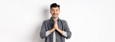 Lütfen bana yardım et. Gülümseyen sevimli adam namaste jestinde el ele tutuşuyor, iyilik istiyor, teşekkür ediyor, umut dolu bakıyor ve kameraya heyecanlı bakıyor, beyaz arka planda duruyor..