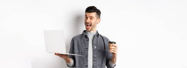 Kahve içen ve dizüstü bilgisayarla çalışan mutlu erkek işçi, kameraya göz kırpıyor, beyaz arka planda duruyor..