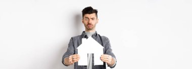 Gayrimenkul ve sigorta konsepti. Kafası karışmış adam kağıttan eve bakıyor. Emin değil, kaşları çatık, beyaz arka planda duruyor..