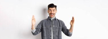 Bıyıklı mutlu bir adam gururlu bir gülümseme ile büyük bir nesne şekillendiriyor, beyaz arka planda ceketle duruyor..