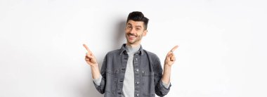 Bıyıklı genç yakışıklı adam iki logo gösteriyor, kameraya mutlu bir şekilde gülümsüyor, beyaz arka planda günlük giysiler içinde duruyor..