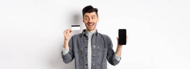 Plastik kredi kartı ve boş akıllı telefon ekranı, reklam uygulaması, beyaz arka planda duran heyecanlı yakışıklı adam..