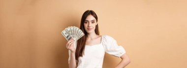 Dolarları gösteren, para kazanan ve bej arka planda nakit parayla duran kendine güvenen, güler yüzlü bir kadın..