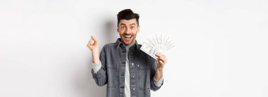 Neşeli genç adam heyecandan zıplıyor ve dolarları gösteriyor, ödül parası kazanıyor, para kazanıyor ve seviniyor, beyaz arka planda duruyor..