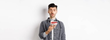 Yakışıklı doğum günü çocuğu pastaya mum üflüyor ve dilek tutuyor, kameraya bakıp, beyaz arka planda duruyor..