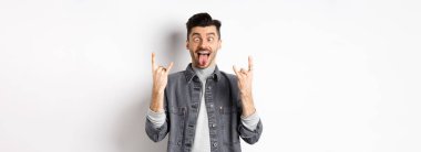 Dilini ve kaya boynuzlarını gösteren komik genç adam, eğlenmek için gözlerini kısarak bakan, parti ya da konserin tadını çıkaran, beyaz arka planda duran.