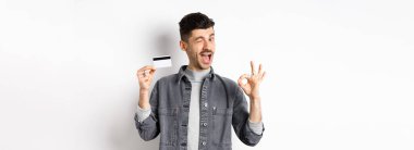 - Çok iyi. Plastik kredi kartı ile gülümseyen adam tamam işareti gösteriyor, gülümsüyor ve göz kırpıyor, bankayı tavsiye ediyor, beyaz arka planda duruyor..