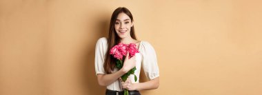 Sevgililer Günü. Romantik kız kameraya mutlu bir şekilde gülümsüyor, sevgilisinden bir buket pembe gül alıyor, bej arka planda duruyor..