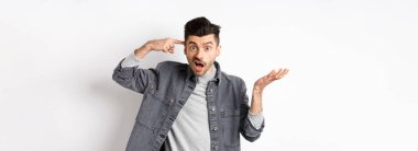 Delirdin mi sen? Sinirli bir adam aptalca davranışları için insanları azarlıyor, baş tapınağını işaret ediyor ve kafası karışmış bir şekilde elini kaldırıyor, beyaz arka planda dururken şikayet ediyor..