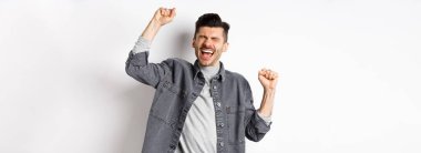 Başarının tatlı tadı. Mutlu genç adam evet diye bağırıyor, mutlu ve rahatlamış yüzüyle yumruk tokuşturuyor, amacına ulaşıyor ve kutluyor, kazanan ödül olarak zafer kazanıyor, beyaz arka plan.