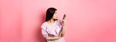 Asyalı kız akıllı telefon ekranına şaşırmış görünüyor, mesaj okuyor, pembe arka planda giyinmiş duruyor..