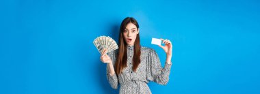 Heyecanlı şanslı kız plastik kredi kartı ve para gösteriyor, hayret içinde, pahalı bir şey alıyor, mavi arka planda duruyor..