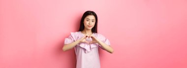 Sevgililer Günü konsepti. Güzel Japon genç kız kalp jesti ve gülümseme şirin, aşığına sevgi ve sempati göster, pembe arka planda romantik elbiseler içinde duruyor..