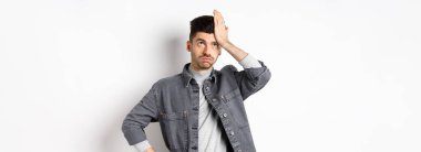 Sinirli genç adam yorgun ya da rahatsız görünüyor, gözlerini yuvarlıyor ve alnını tokatlıyor, aptalca bir şeyden facepalm yapıyor, beyaz arka planda duruyor..