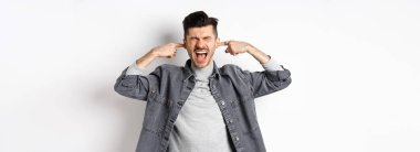 Sinirli adam kulaklarını kapattı ve çığlık attı, yüksek sesten rahatsız oldu, gürültülü müziği durdurmak için bağırdı, beyaz arka planda durdu..