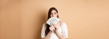 Dolarları tutan ve boş alana bakan düşünceli bir kız. Parayla ne alacağını düşünüyor, alışverişi planlıyor, bej arka planda duruyor..