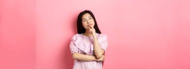 Dalgın gülümseyen Asyalı kız seçim yapıyor, sol üst köşedeki logoya bakıyor ve düşünüyor, bir fikri var, pembe arka planda kurnazca duruyor..