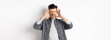 Sinirli genç adam bağırıyor ve kafaya dokunuyor, ayakta duruyor gürültüden ya da baş ağrısından rahatsız oluyor, beyaz arka planda duruyor..
