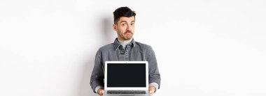 Belirsiz genç adam somurtuyor ve omuz silkiyor boş dizüstü bilgisayar ekranı gösterirken, beyaz arka planda habersiz duruyor.