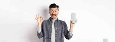 Heyecanlı gülümseyen adam boş akıllı telefon ekranı gösteriyor. İyi bir jest yapıyor, uygulama veya alışveriş öneriyor, beyaz arka planda duruyor..