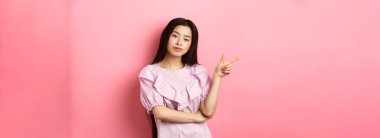 Genç kendini beğenmiş Asyalı kız havalı görünüyor ve logoyu işaret ediyor pembe romantik arka planda reklam ürünü..