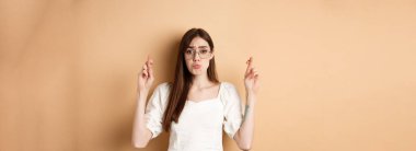 Dilek tutan, dudaklarını büzen ve yalvaran tatlı, umut dolu kız, lütfen söyle, bej arka planda duruyor..