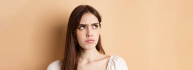 Kızgın, sevimli, somurtkan, canı sıkkın, kötü şeylere bakan, bej arka planda duran.