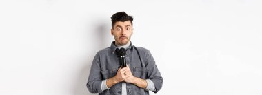 Gergin, kendine güveni olmayan, mikrofon tutan, kararsız görünen, gösteri yapmaya korkan, beyaz arka plana karşı utangaç bir adam..